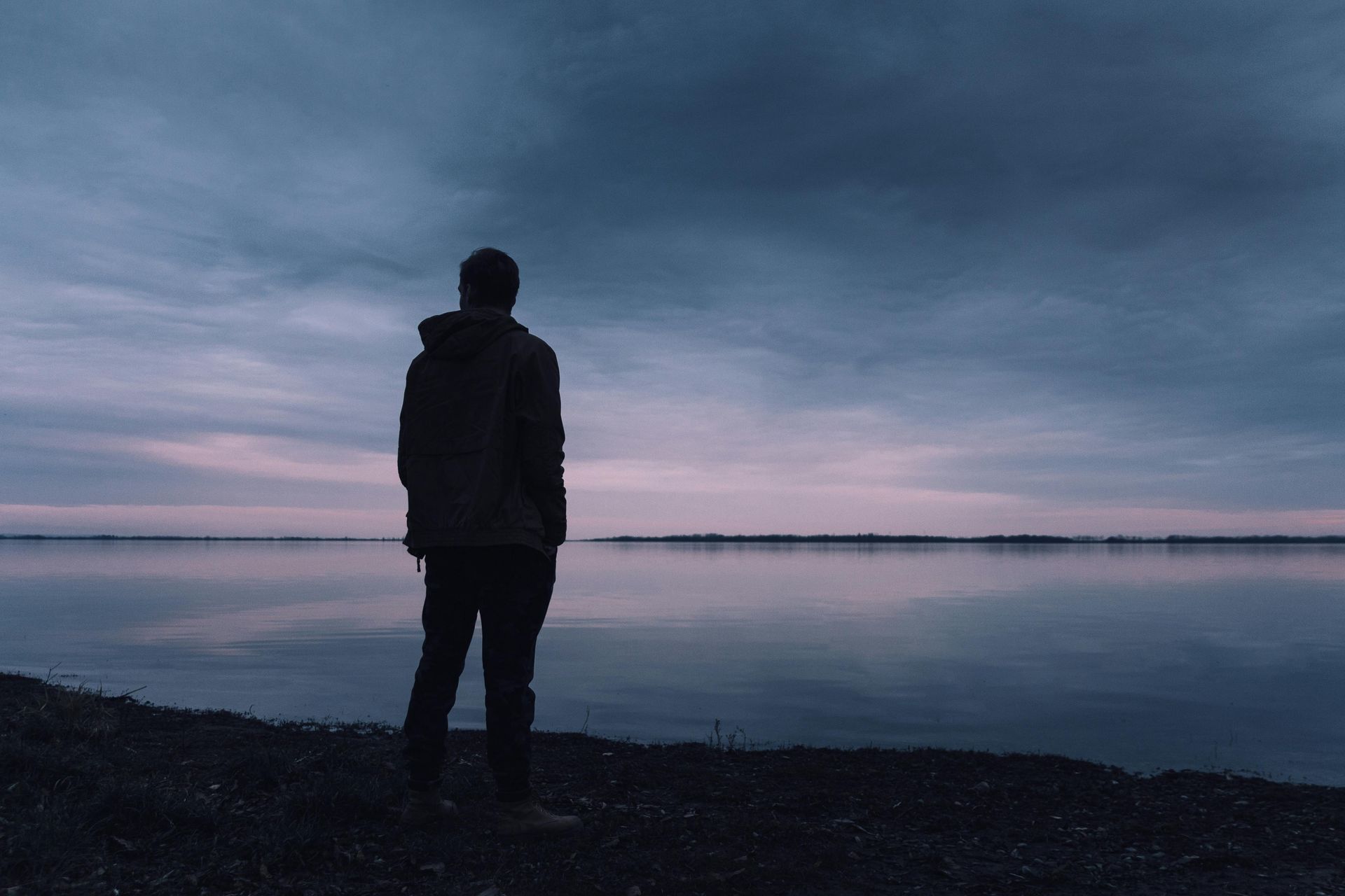 Person standing by calm water, looking at a dusky sky. Reflections of the sky in the water.
