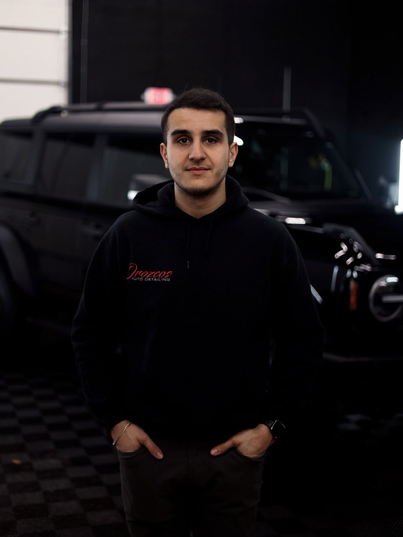 A man in a black hoodie is standing in front of a black car.