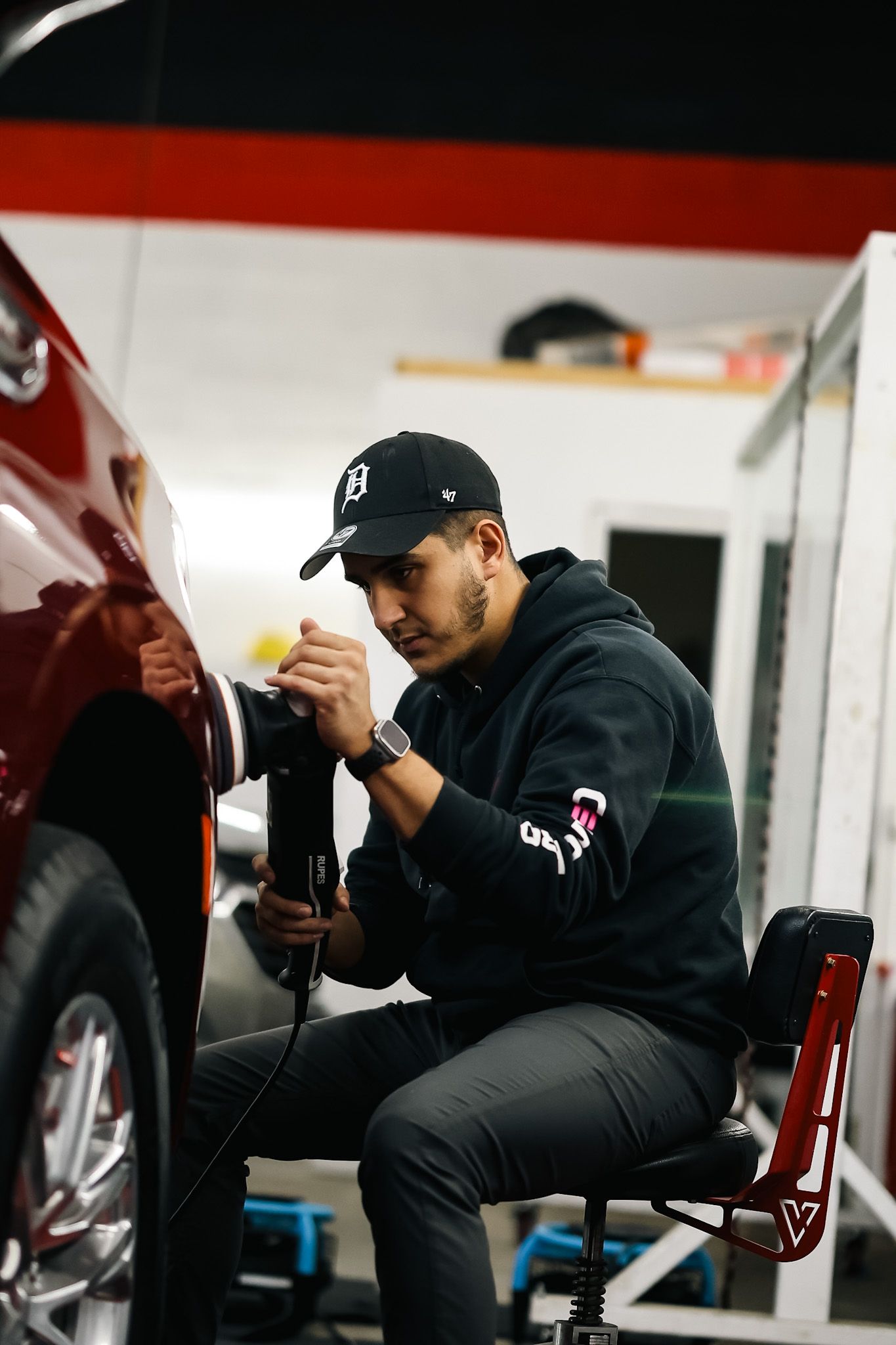 Mechanic working on a red car, using a tool while sitting in a chair, inside a garage.