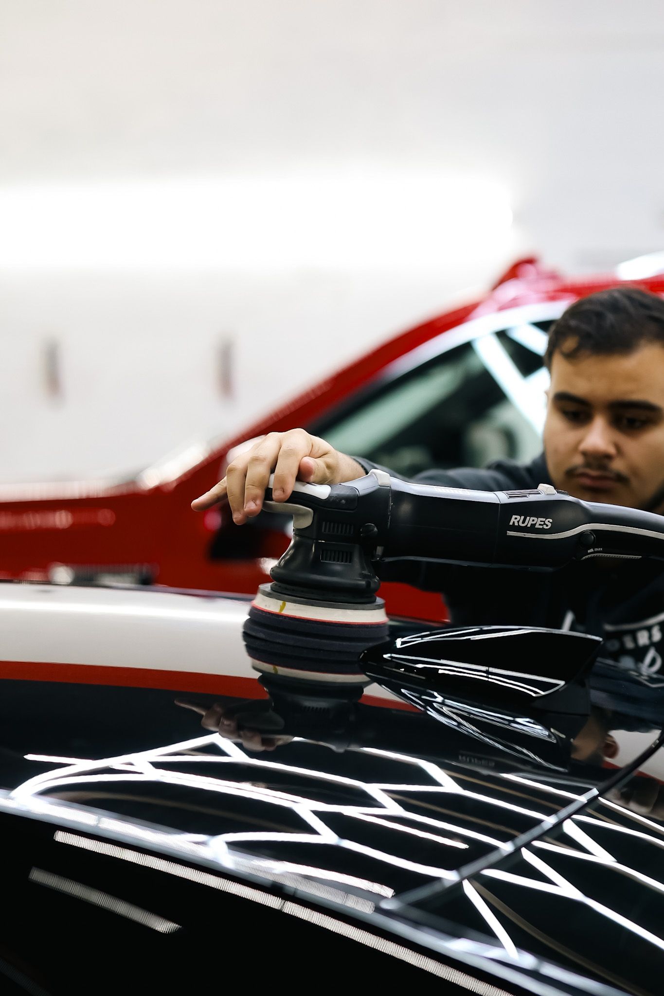 Man polishing a black car with a rotary buffer in a garage; red car in background.