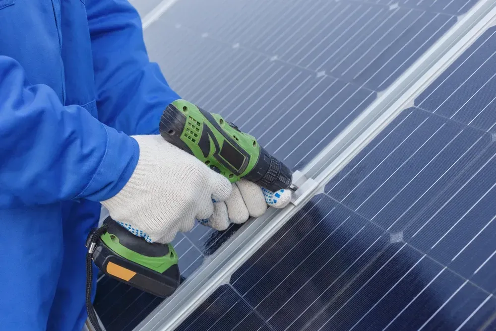 Person in blue coveralls installing solar panel with a power drill. — OzEnergi in Pimpama, QLD