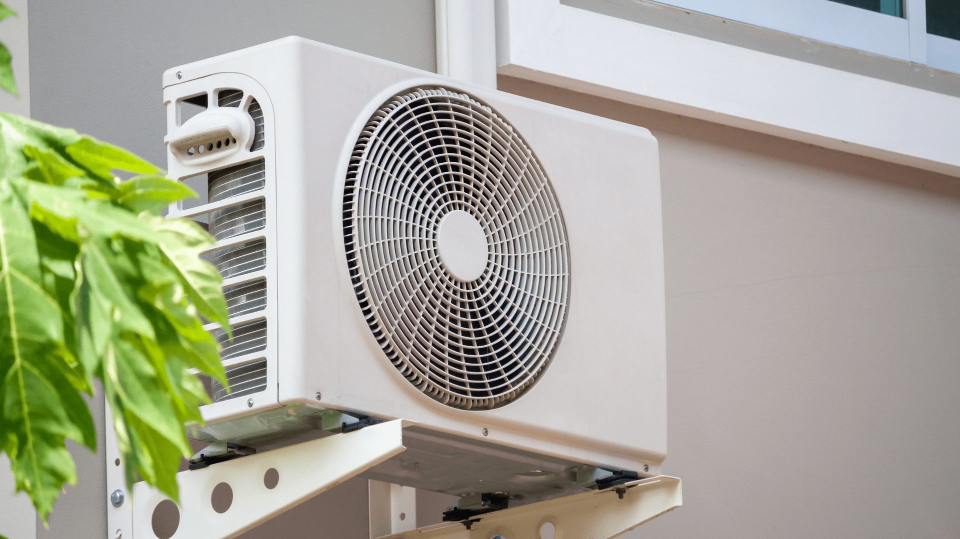 White outdoor air conditioning unit mounted on a light gray wall, with green foliage to the left — OzEnergi in Caloundra West, QLD