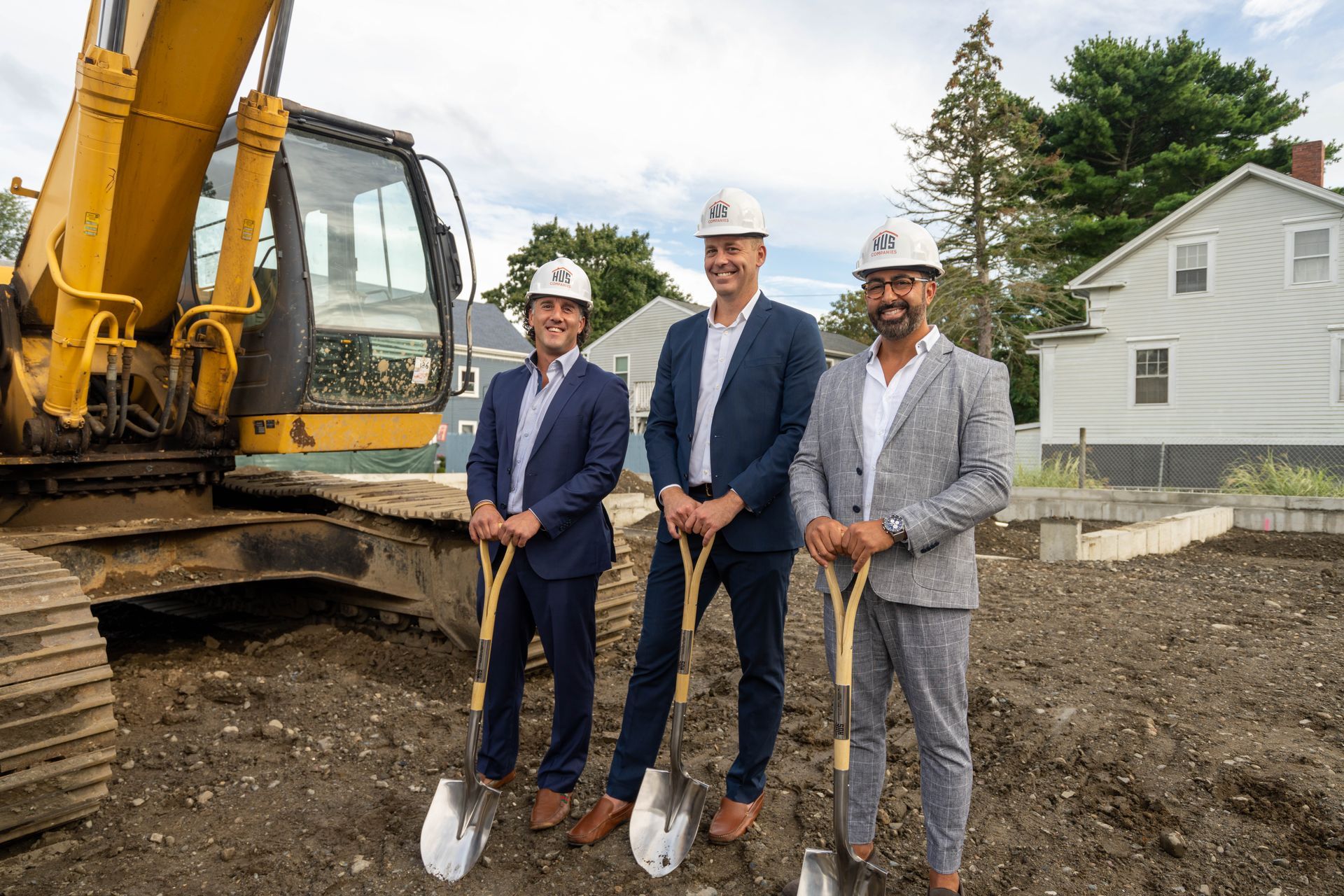 Three men in suits and hard hats are holding shovels in front of a construction vehicle.