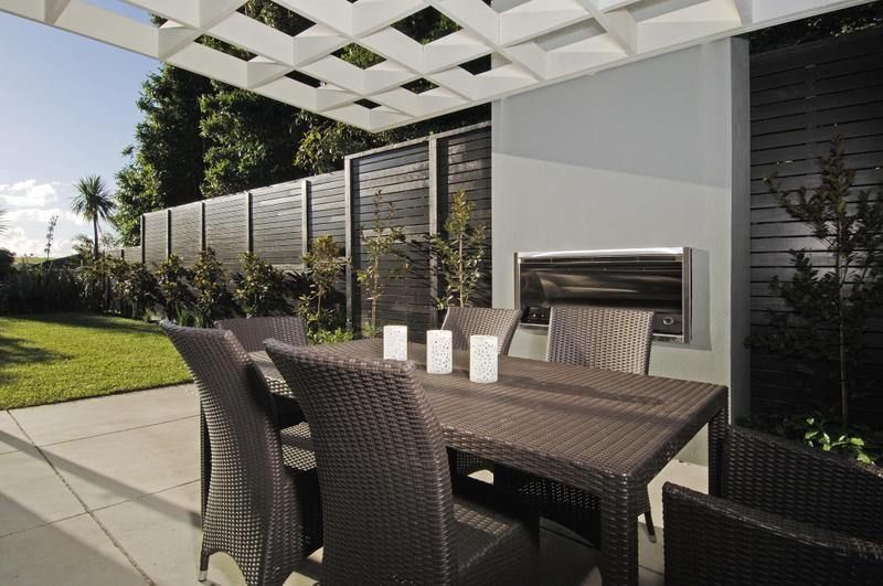Outdoor dining area with a table and chairs under a latticed pergola. Grill and fence in background.
