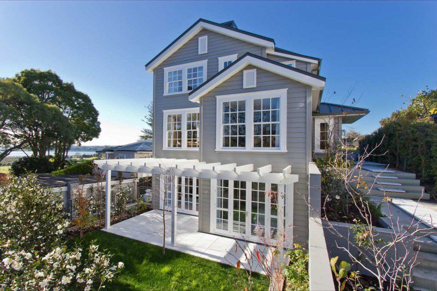 Two-story gray house with white trim and pergola, surrounded by greenery and a sunny blue sky.