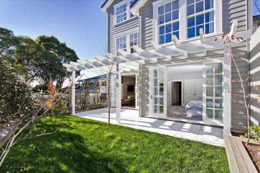 Backyard of a gray house with a white pergola, white-framed French doors, and a green lawn.