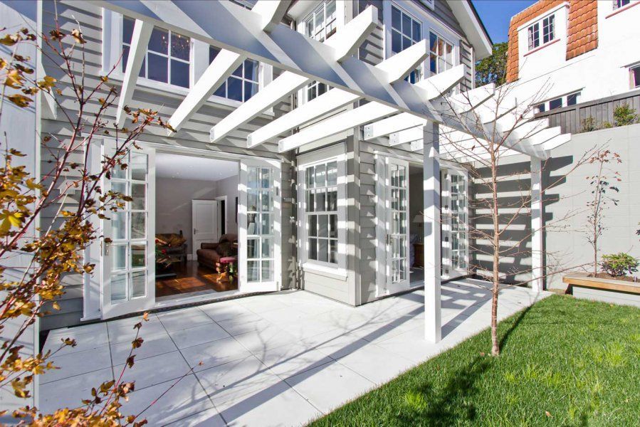 Grey house exterior with white pergola and patio, French doors open to living area, green lawn.