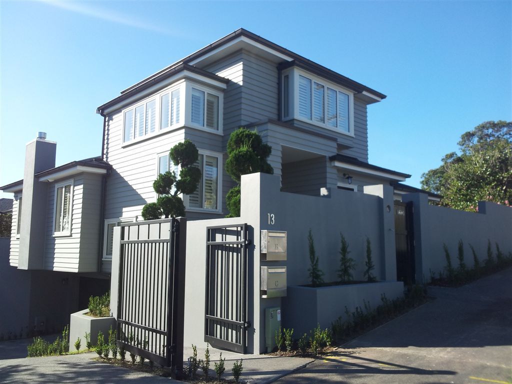 Gray, two-story house with a black gate, mailbox, and a high wall against a blue sky.