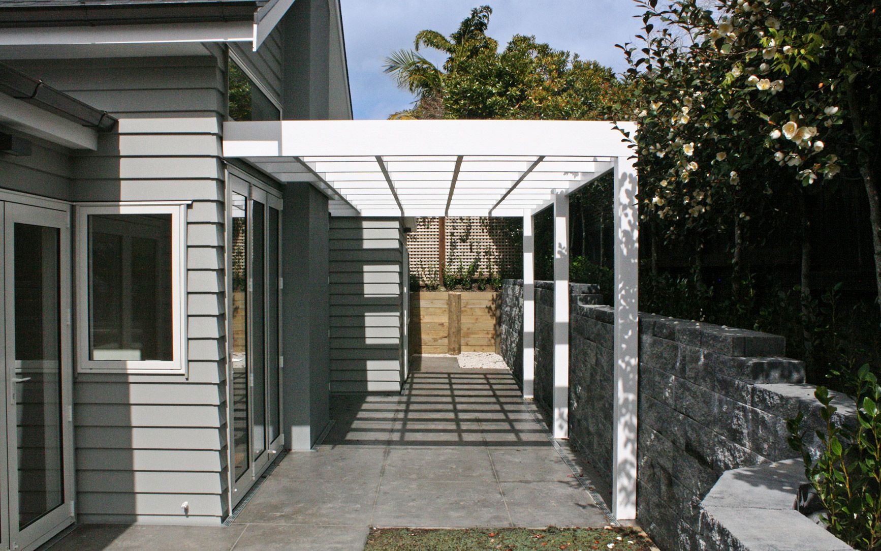 Pergola extending from a gray house, casting shadows on a pathway with a stone wall and landscaping.
