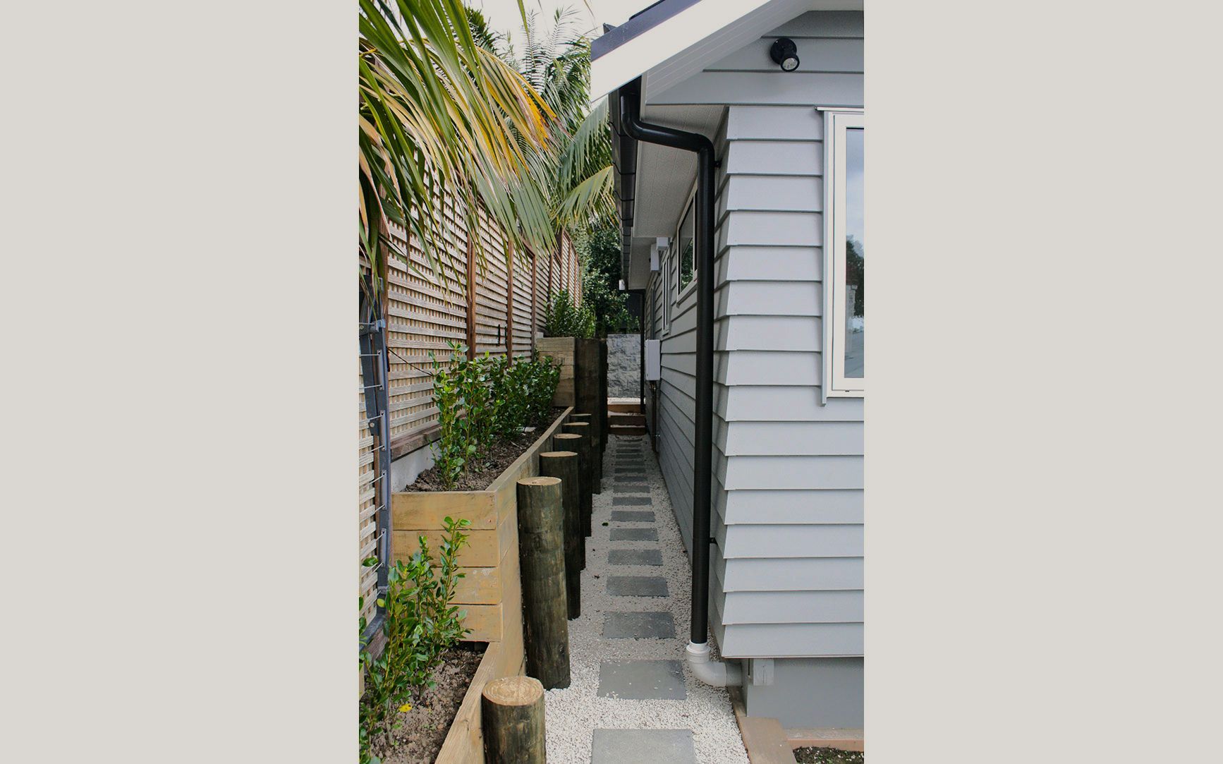 Narrow outdoor walkway between a gray-sided building and a planter with trees, pathway of stepping stones.