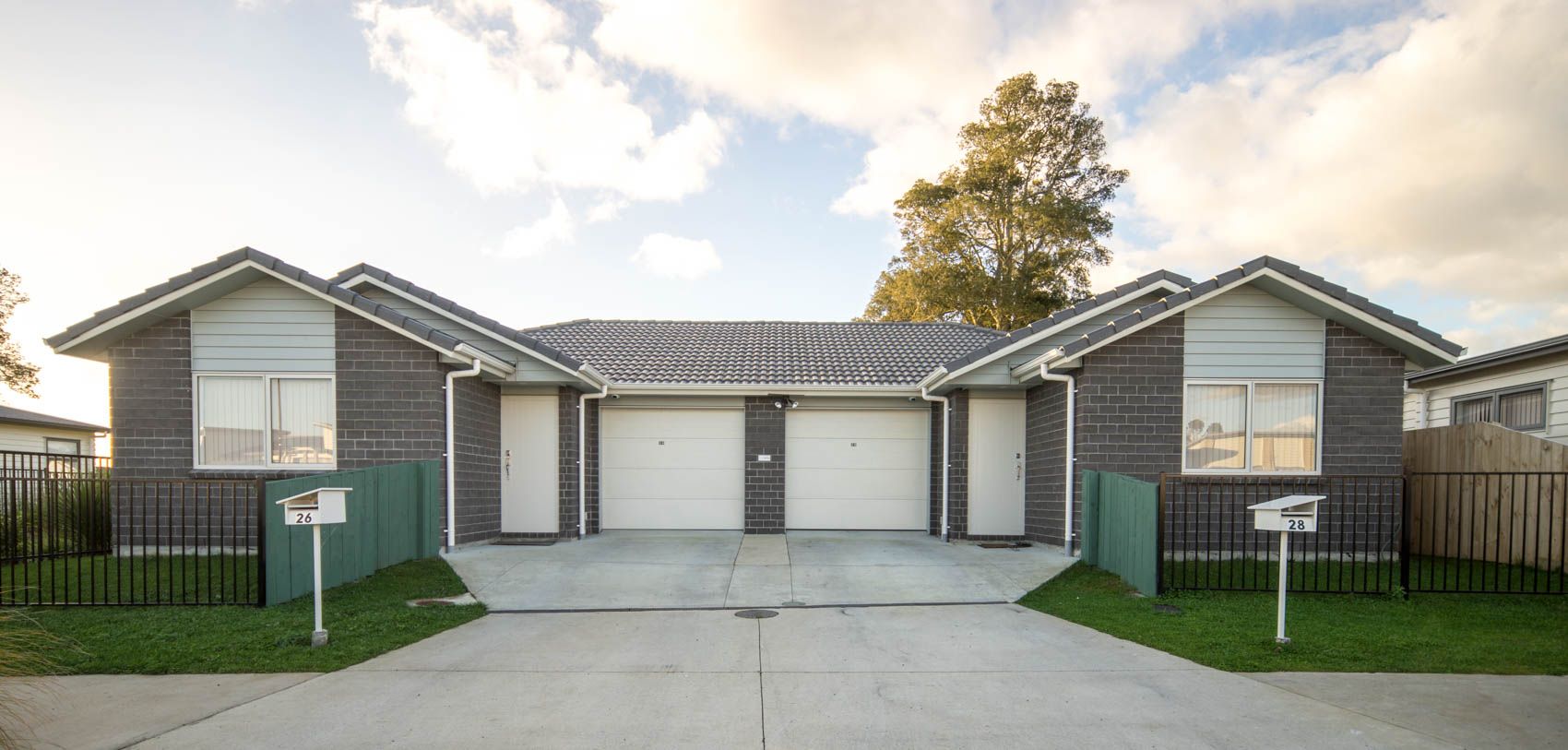 Two-story duplex houses with grey brick facade, white garage doors, and green lawns against a cloudy sky.