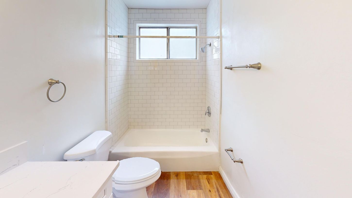 White bathroom with a bathtub, toilet, and window above. Light-colored wood floor.