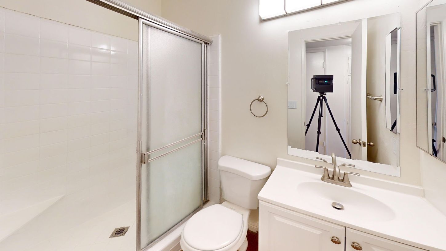 White bathroom with a shower, toilet, sink, and mirror. A camera on a tripod is reflected in the mirror.