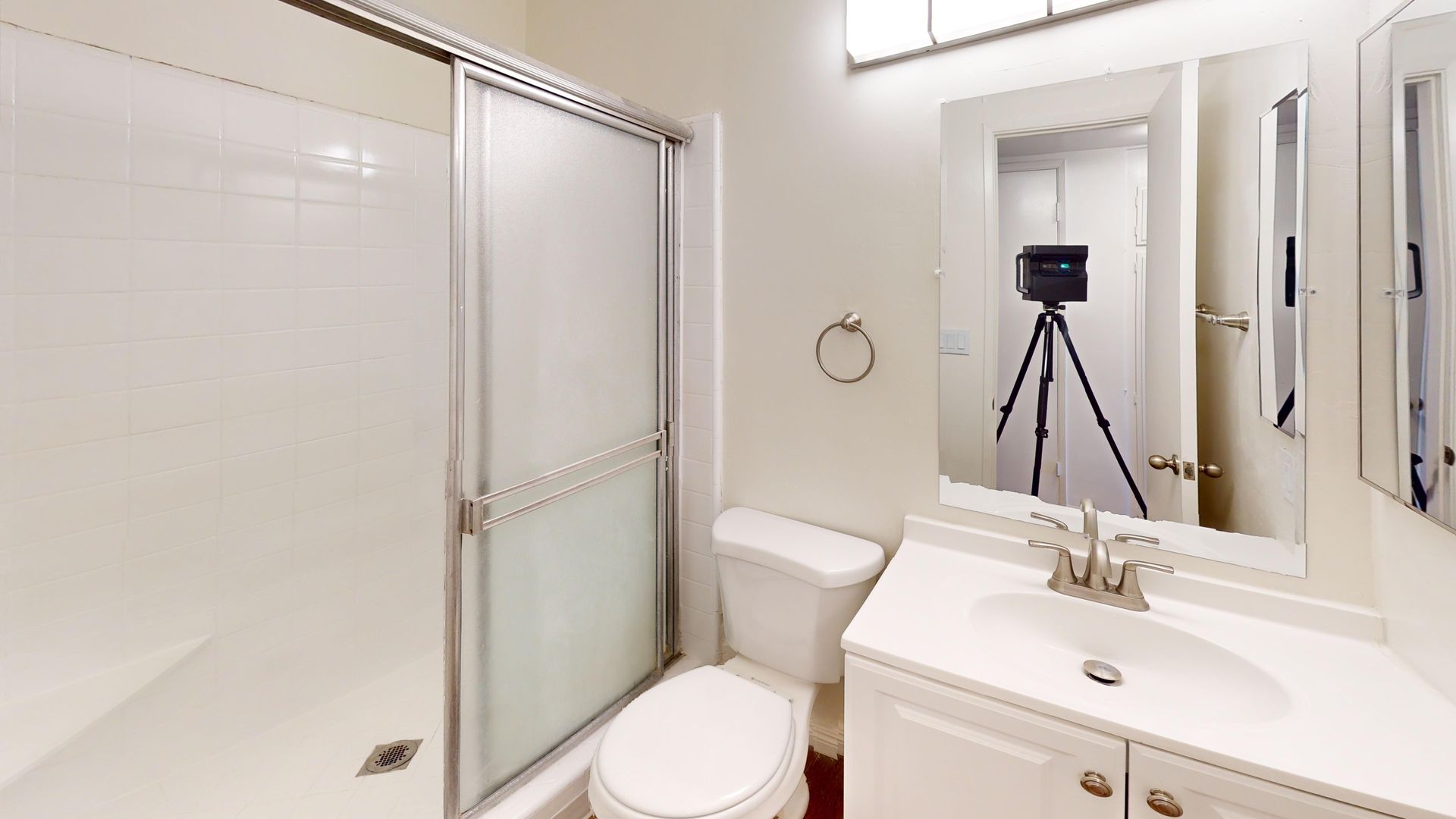 White bathroom with a shower, toilet, sink, and mirror. A camera on a tripod is reflected in the mirror.