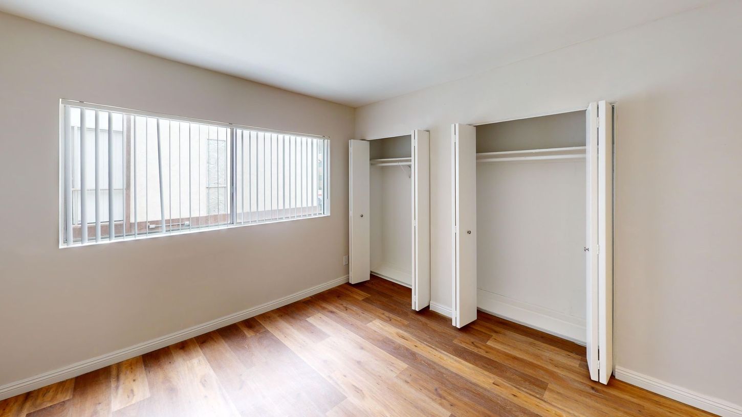 Empty bedroom with hardwood floors, a window with blinds, and two closets.