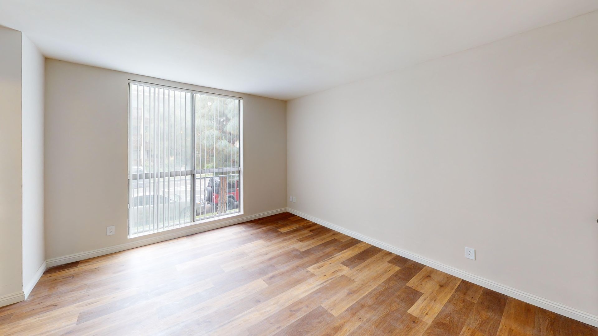 Empty room with wood floor, large window with blinds, white walls.