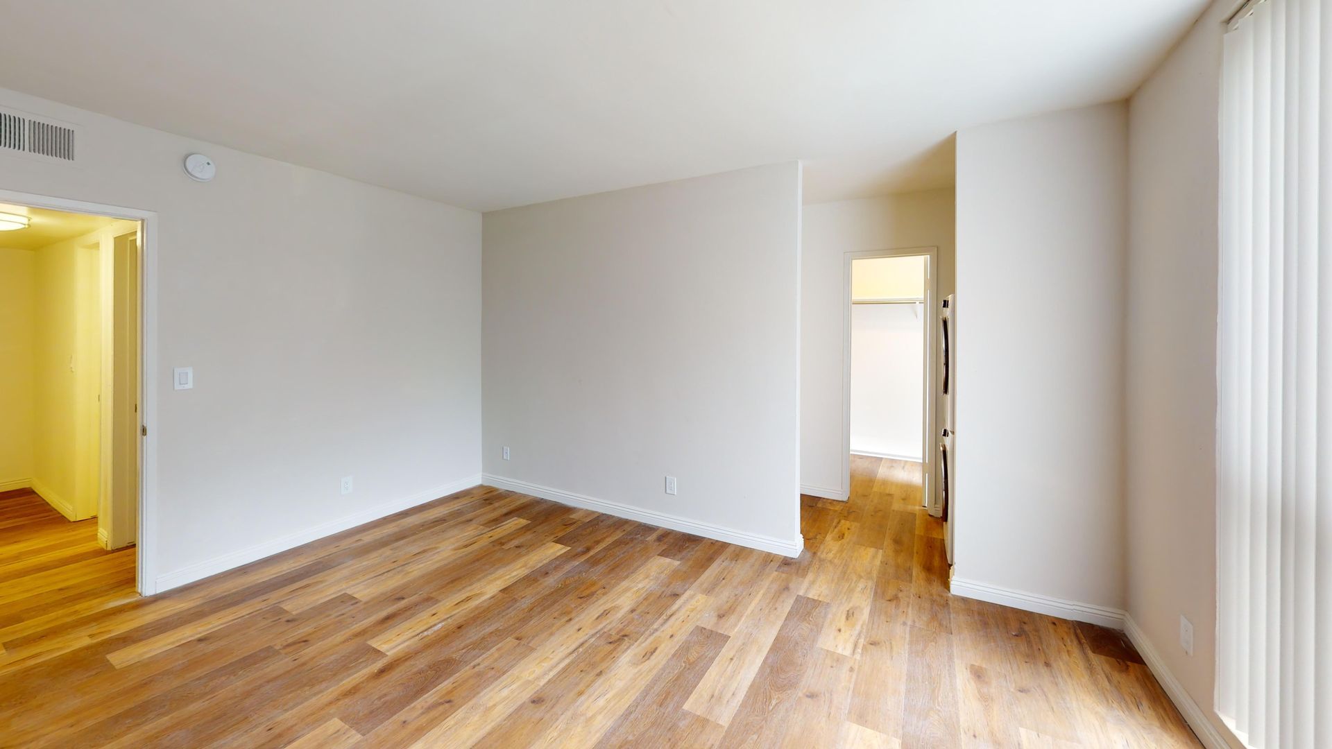 Empty room with wood floors and white walls, door to the left, hallway in the background, and window with blinds.