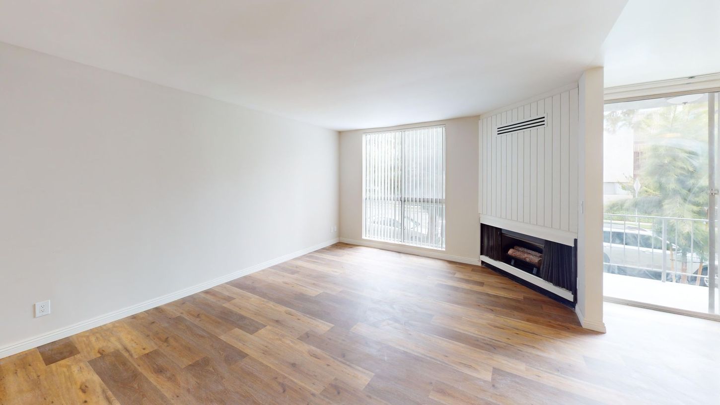 Empty living room with wood floor, white walls, and a fireplace. Large windows.