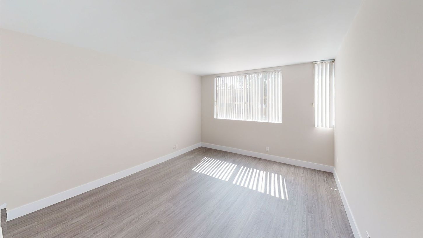 Empty, sunlit room with gray wood-look flooring and cream-colored walls. Two windows with vertical blinds.