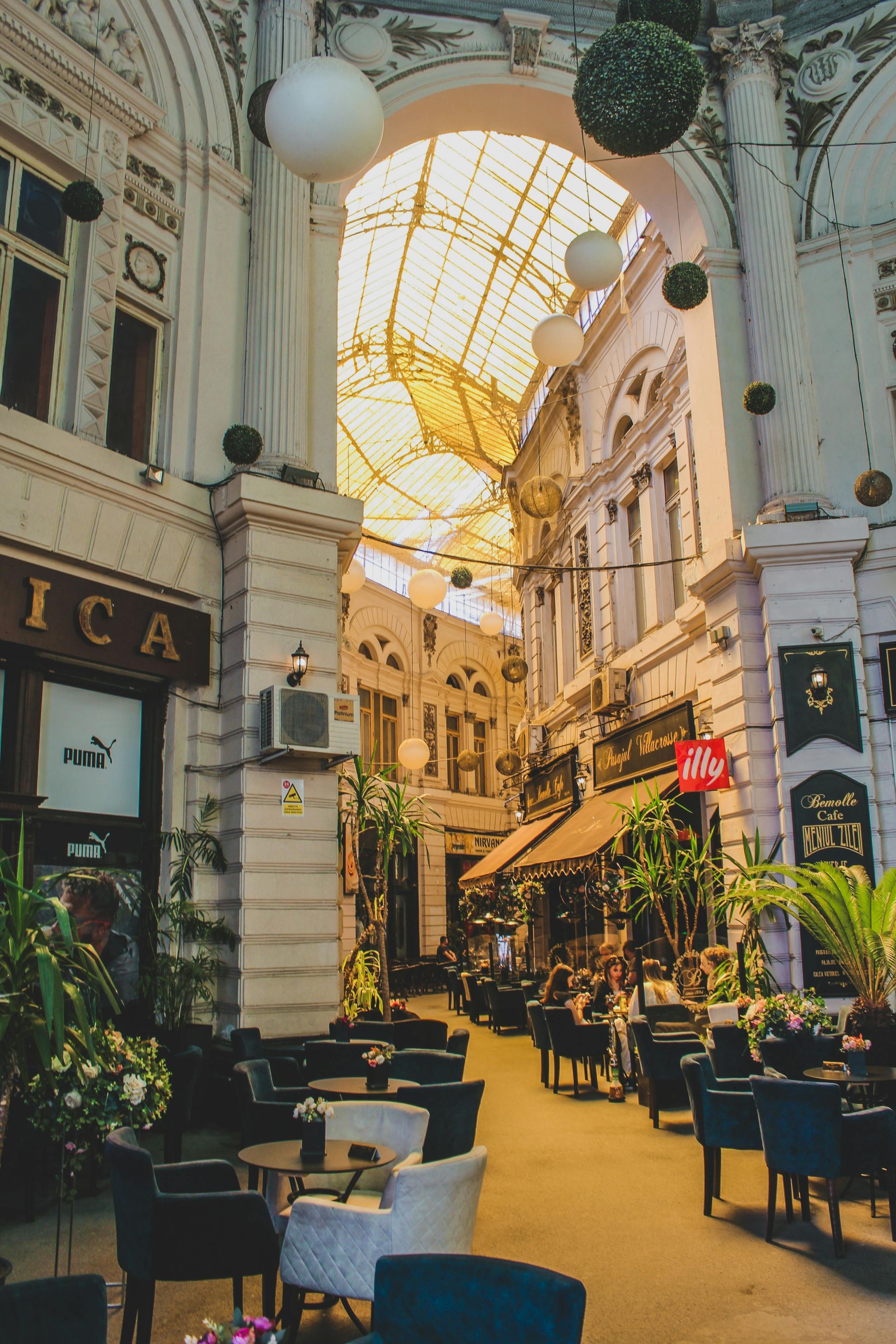 Indoor arcade with cafes and shops under a glass roof. Tables and chairs are set up.