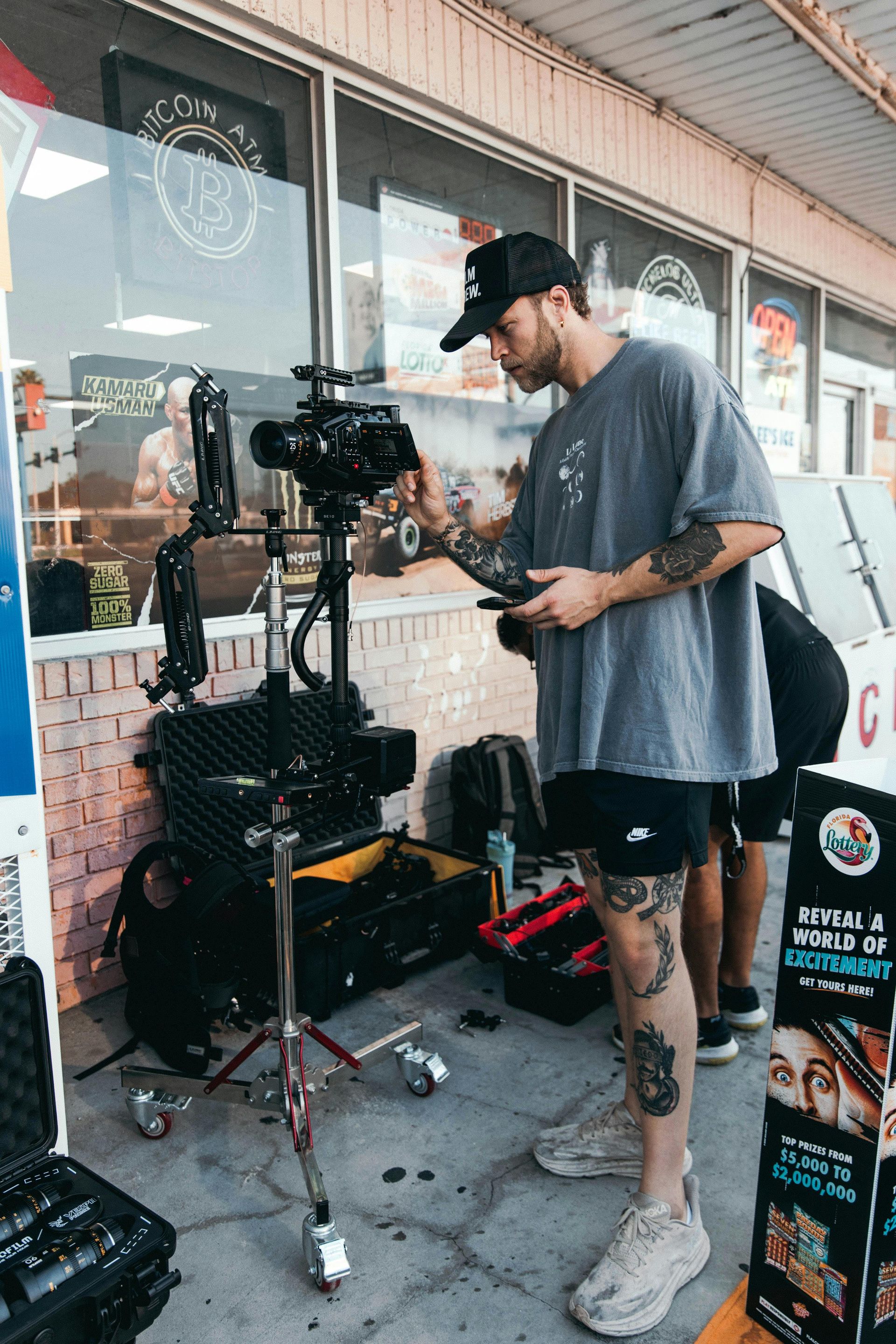 Man operating a large camera setup outdoors. He wears a hat, shorts, and is focused.