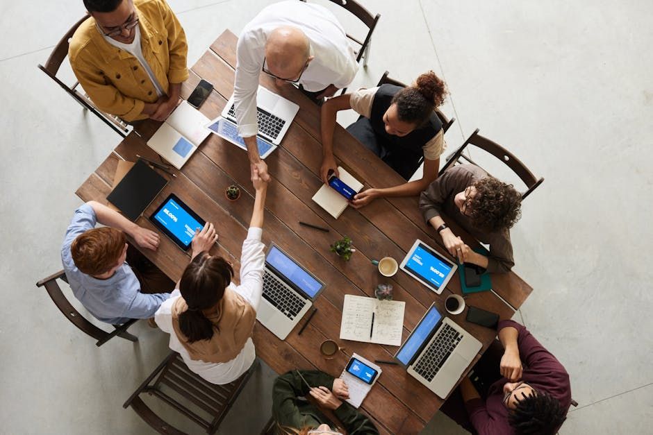 People shaking hands around a wooden table in an office setting, surrounded by laptops, tablets, and notebooks.