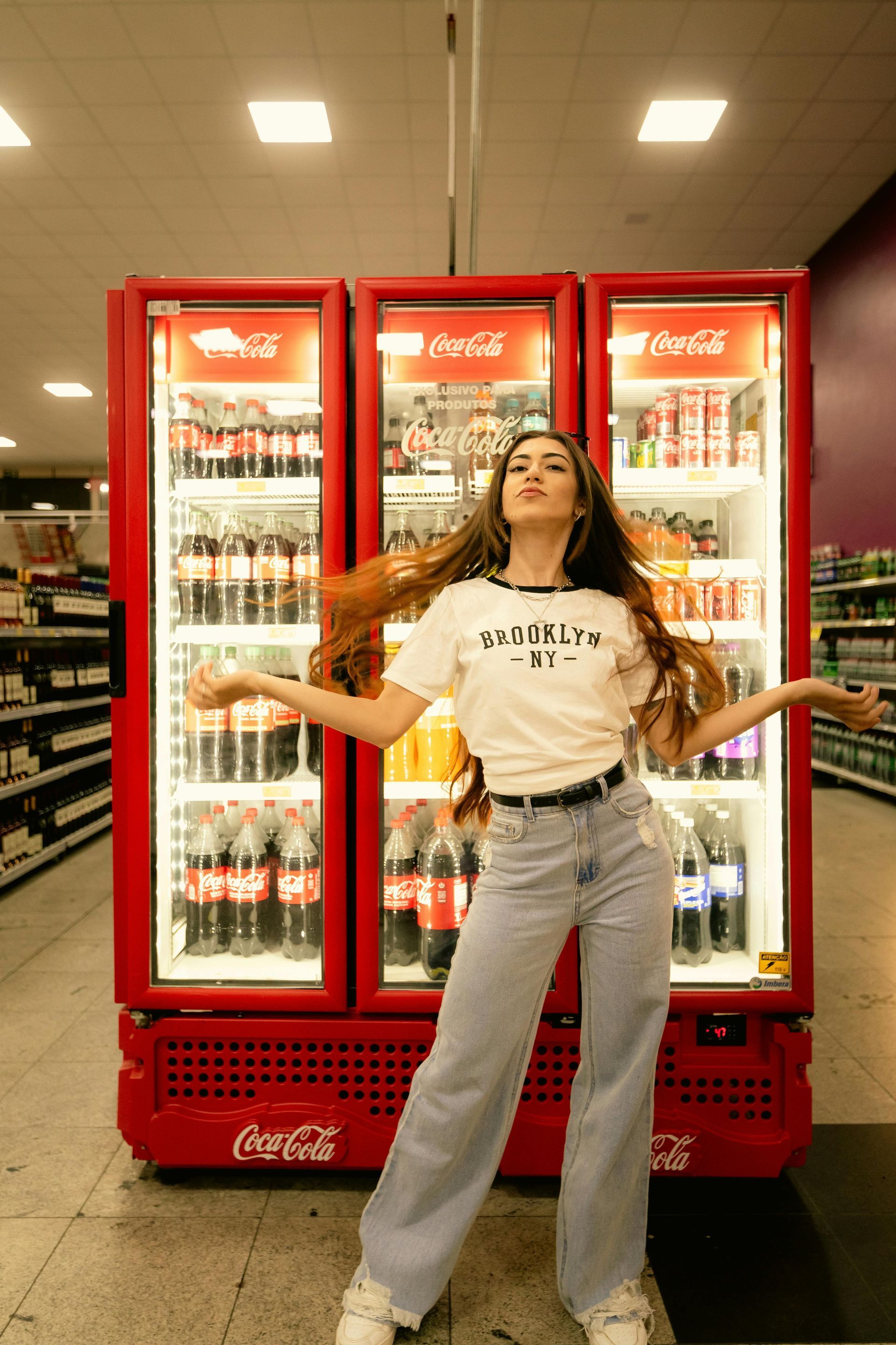 Woman poses in front of Coca-Cola refrigerators in a grocery store, arms outstretched, long hair flowing.