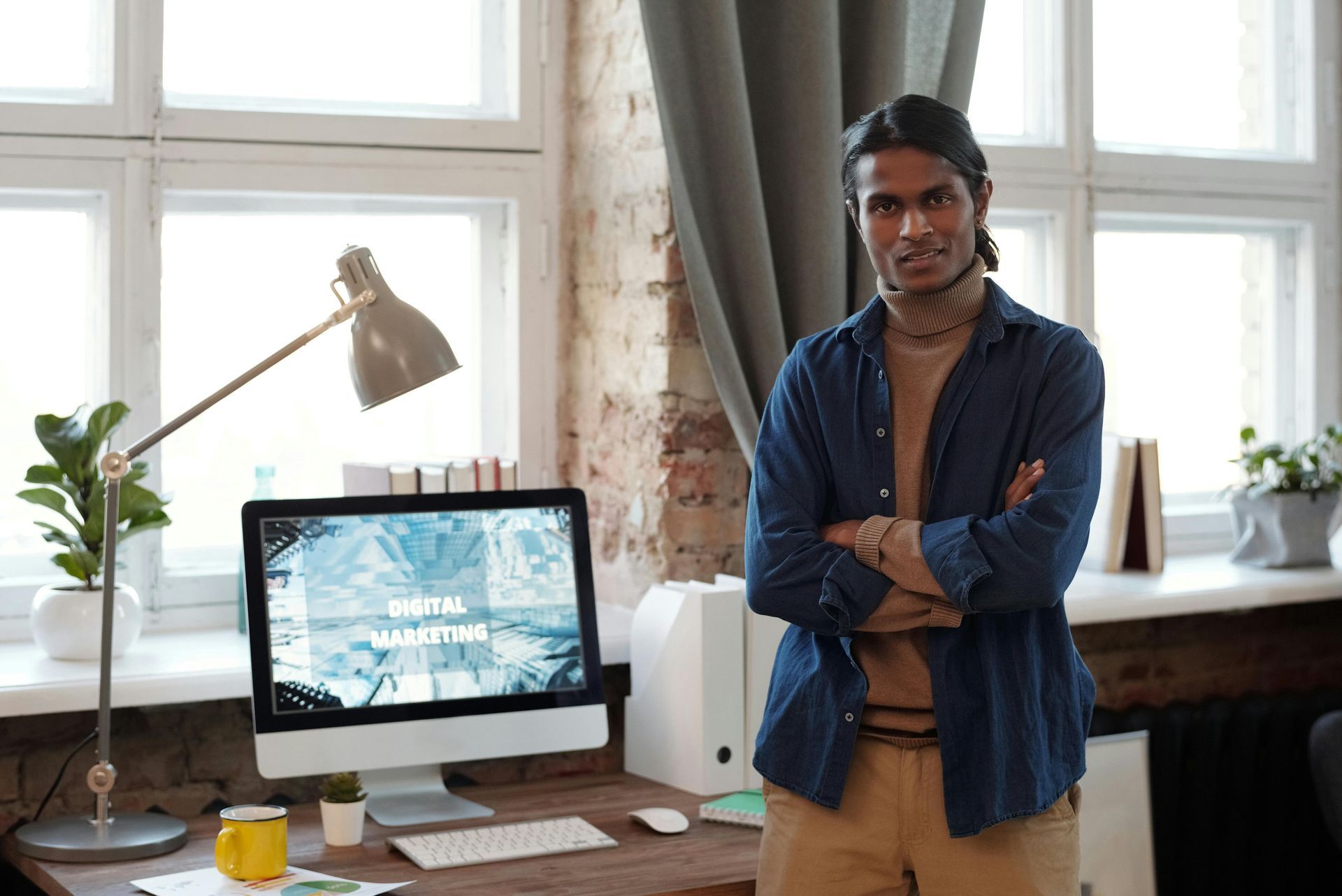 Man with arms crossed standing by desk with computer screen displaying 