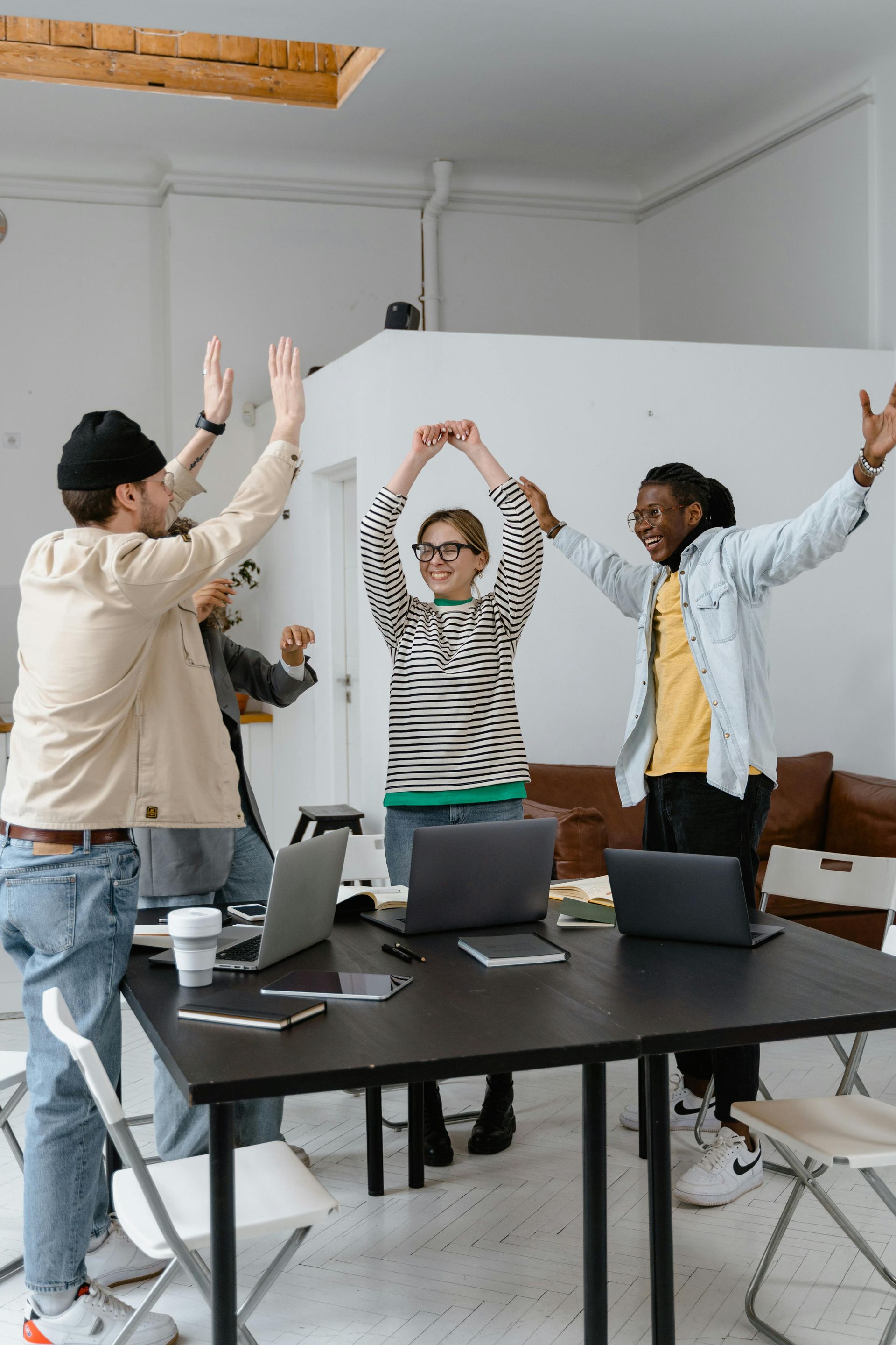 Group of people celebrating around a table with laptops, arms raised in a brightly lit room.