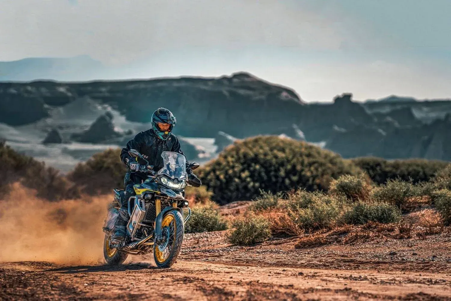 Motociclista pilotando uma moto de trilha em um caminho desértico e empoeirado, com colinas rochosas ao fundo.