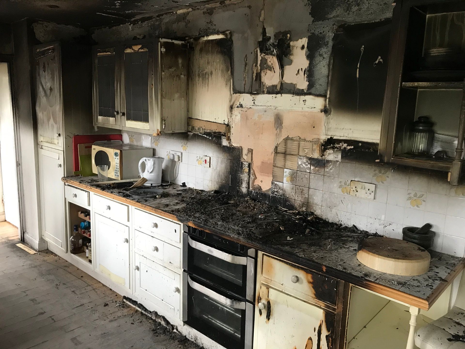 Burnt kitchen interior with charred cabinets, appliances, and countertops. Dark soot covers surfaces.