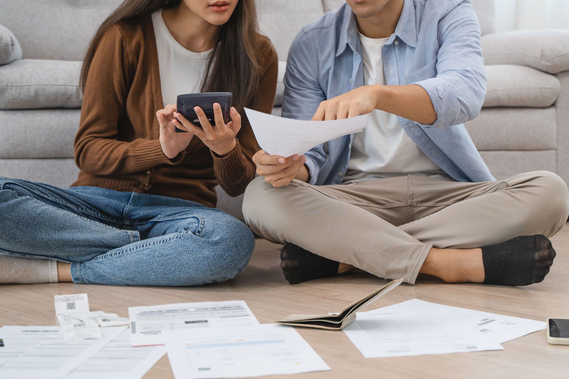 Two people sit on the floor reviewing bills and paperwork, using a phone and calculator in a living room.