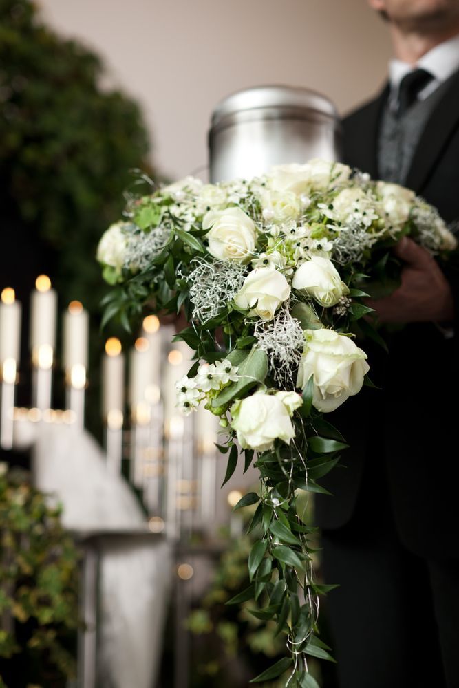 A Man Is Holding A Bouquet Of White Flowers And A Urn At A Funeral — United Funeral Care In Kempsey, NSW