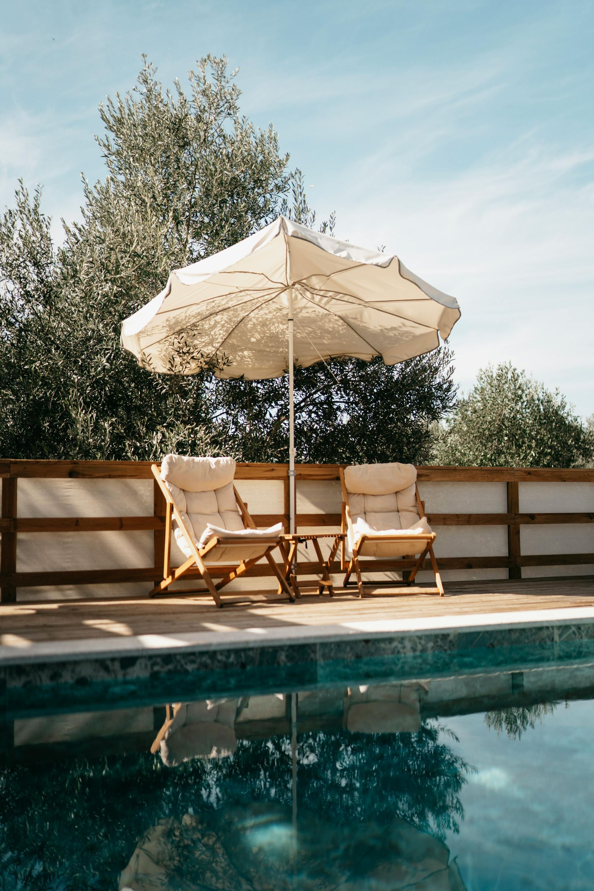 Two beige chairs under a white umbrella by a pool, with trees and a wooden fence in the background.
