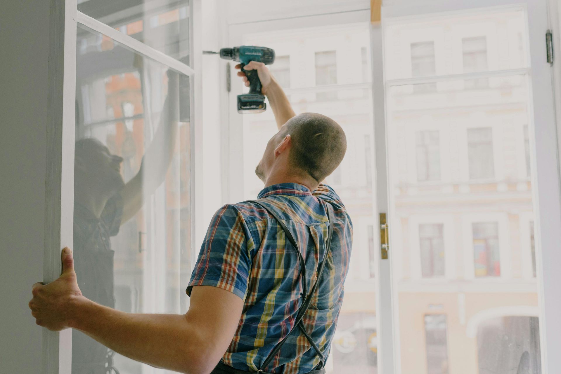 Man in plaid shirt using a drill on a white window frame, with a building in the background.