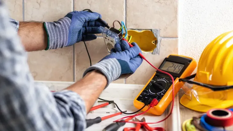 Electrician testing wires in a wall box with a multimeter, blue gloves and hard hat nearby