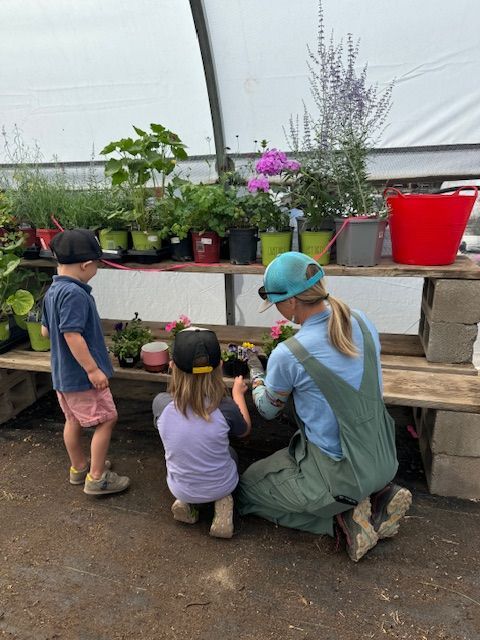 Two children and an adult in a greenhouse tend to small potted plants on a wooden table.