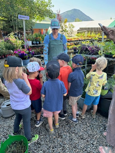 A group of children in hats listens to a person in overalls at an outdoor plant nursery with mountains in the background.