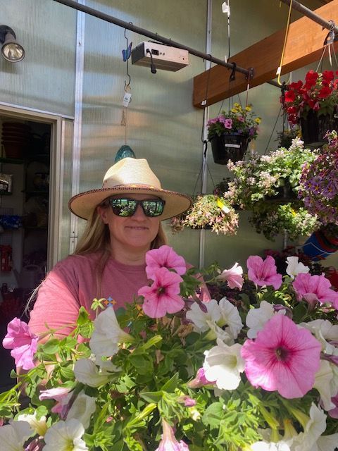 A person wearing a sun hat and sunglasses stands behind hanging flower baskets in a greenhouse.