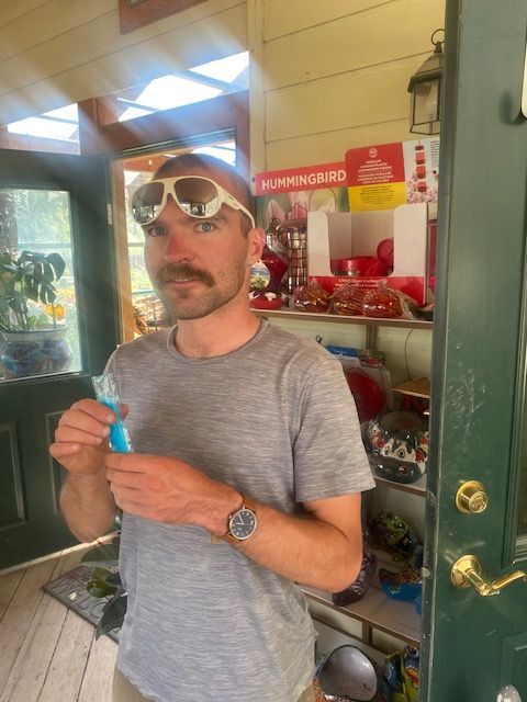 A person with a mustache and sunglasses on their head holds a blue popsicle in a sunlit indoor setting with shelves.