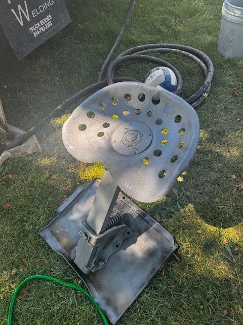 A tractor seat and mounting bracket being spray-painted on grass. Grey paint and green hoses are visible.