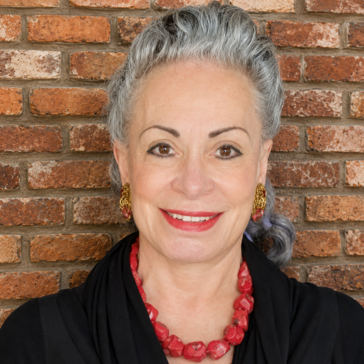 A woman with gray hair and a vibrant red necklace smiles warmly at the camera, radiating charm and confidence.