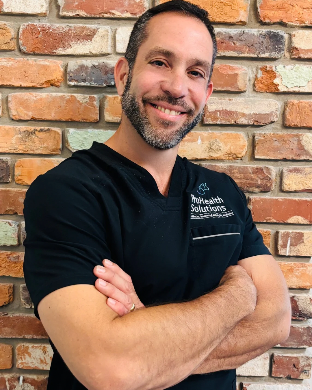 A man with a beard, dressed in a black scrub top, stands with his arms crossed, exuding a calm and professional demeanor.