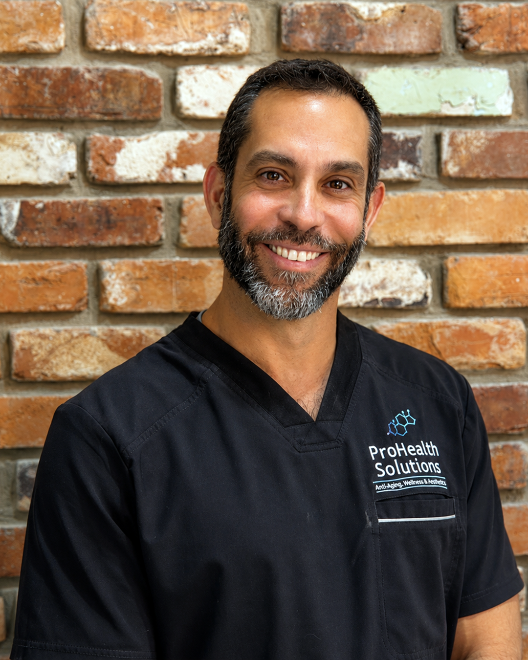 A man with a beard, dressed in a black scrub top, stands with his arms crossed, exuding a calm and professional demeanor.