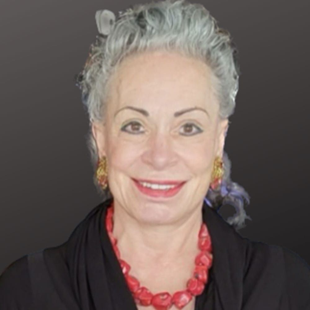 A woman with gray hair and a vibrant red necklace smiles warmly at the camera, radiating charm and confidence.