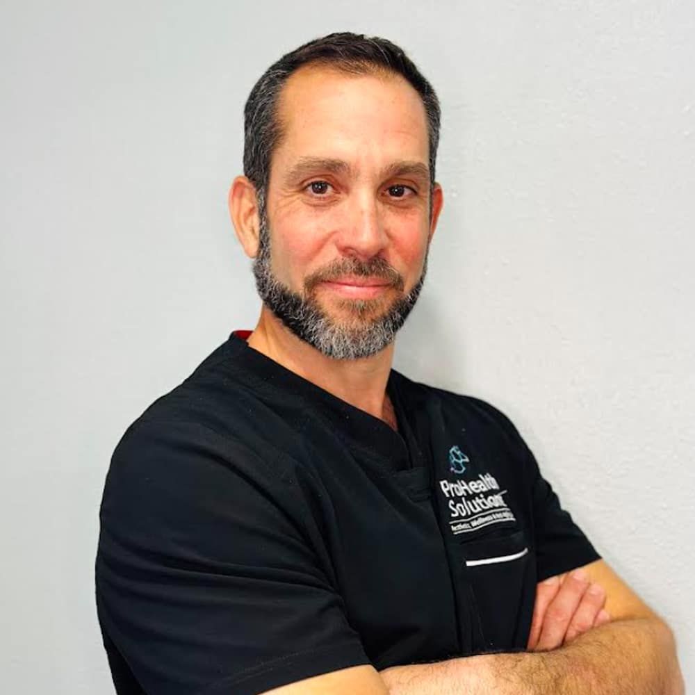 A man with a beard, dressed in a black scrub top, stands with his arms crossed, exuding a calm and professional demeanor.