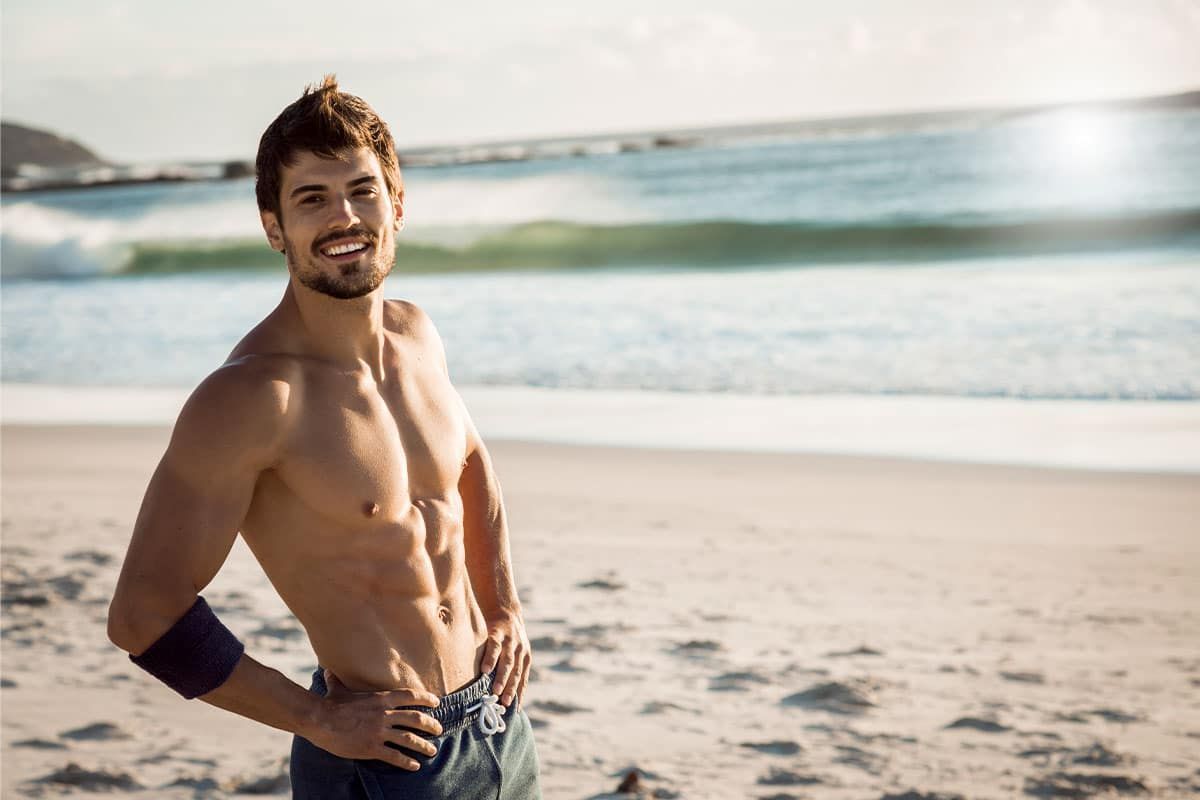 A shirtless man is standing on the beach with his hands on his hips.