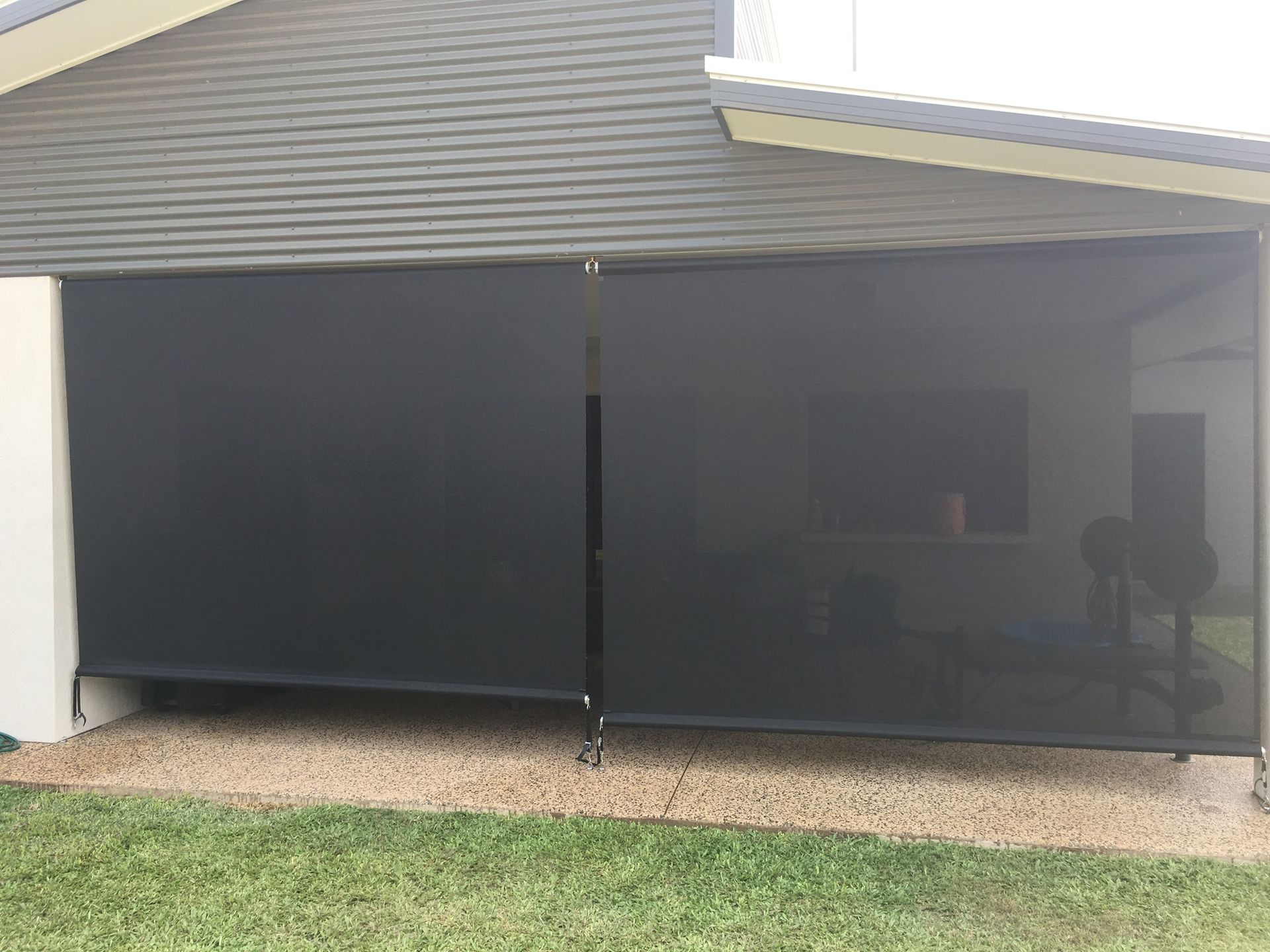 Black outdoor roller shades attached to a building, partially obscuring the interior, on a gravel base next to grass  — NT Blinds in Darwin City, NT