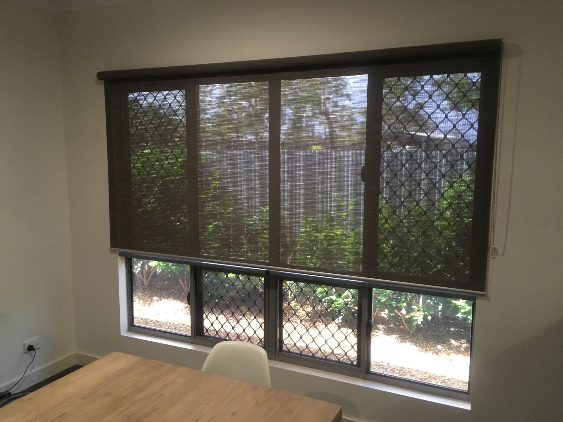Dark roller blind over a window with security screen; interior shot with a glimpse of greenery — NT Blinds in Palmerston, NT