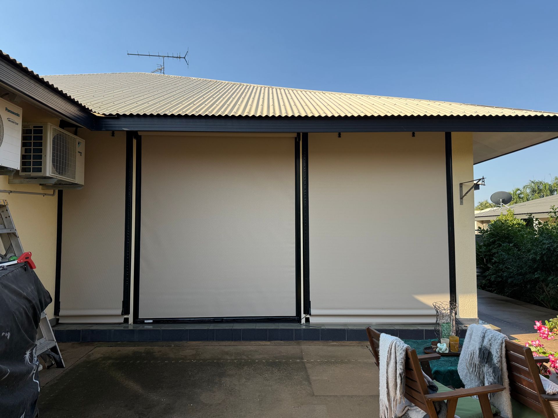 Tan roll-down shades on a house patio. Beige walls, black frames, and a light-coloured roof. Sunny outdoor setting — NT Blinds in Yarrawonga, NT